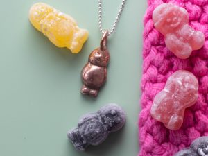 Copper clay jelly baby pendant sat with some jelly babies on a pink and green background.