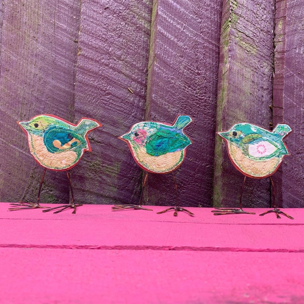 Three green wrens on a pink table by a purple fence